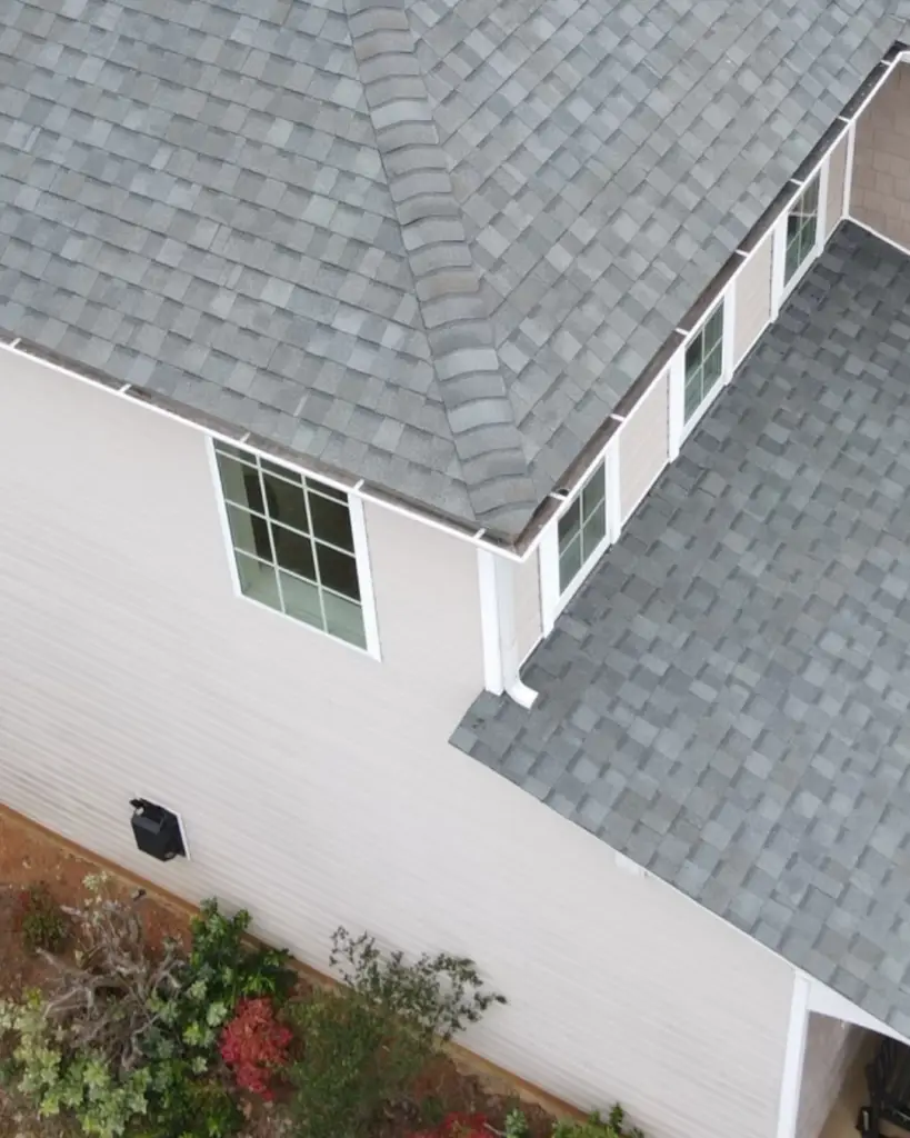 Aerial close-up of a gray shingle roof valley meeting beige siding, with windows, white gutters, and landscaping below.