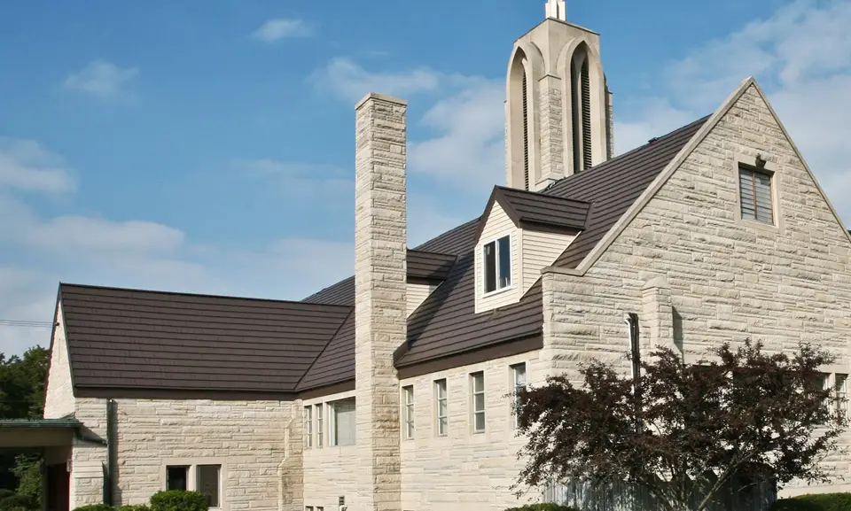 View of a stone church with a dark brown standing seam metal roof, tall vertical steeple, and chimney against a clear sky.