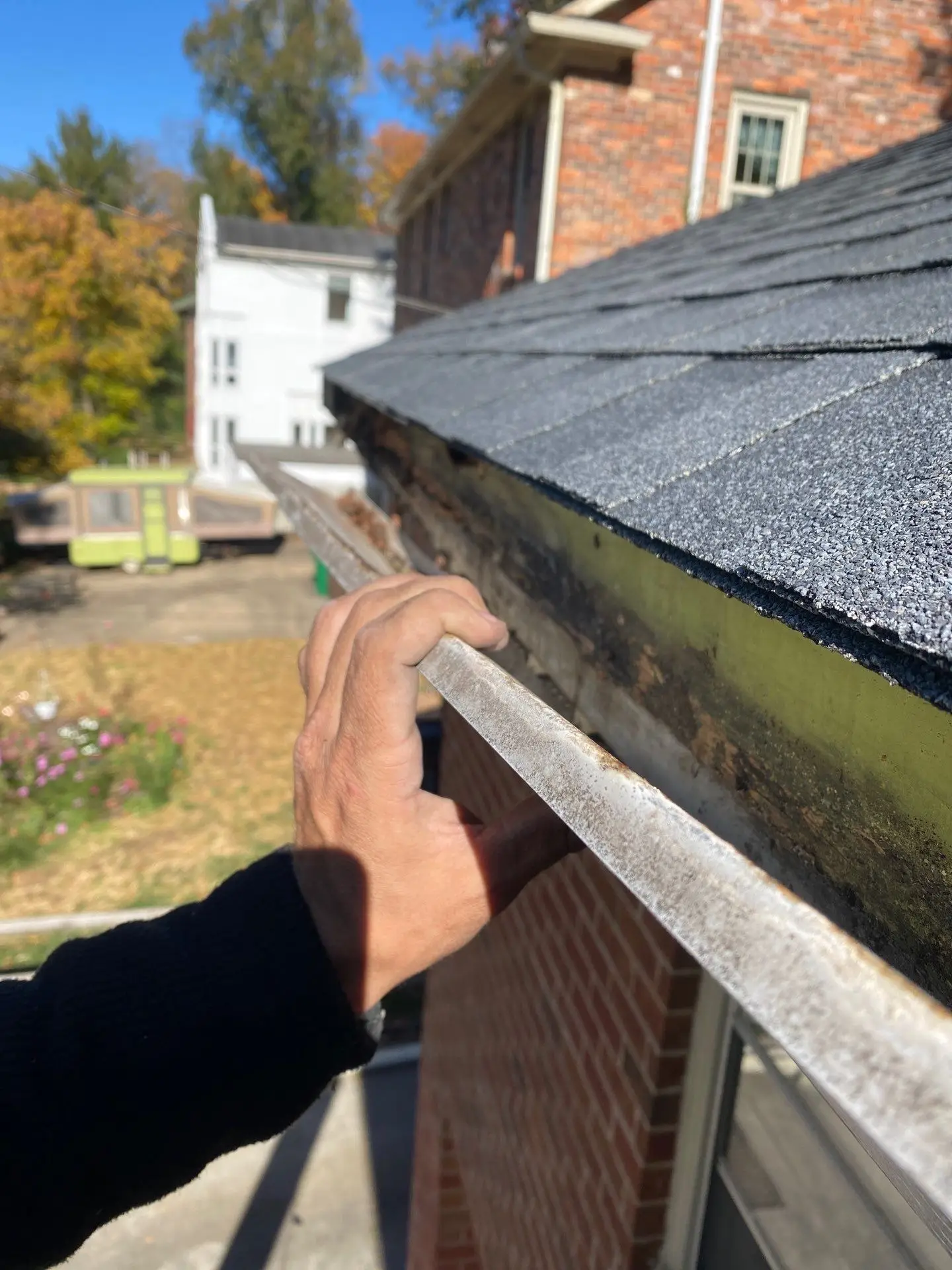 Hand pulling a gutter away from the roof edge to inspect water-damaged fascia beneath asphalt shingles.
