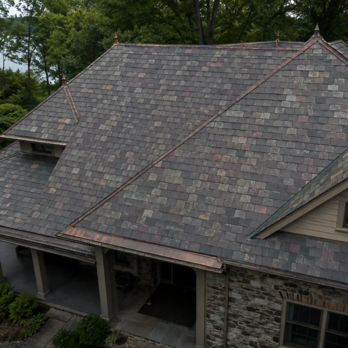 Aerial view of a slate roof with mixed gray tones and copper valley flashing on a stone house surrounded by trees