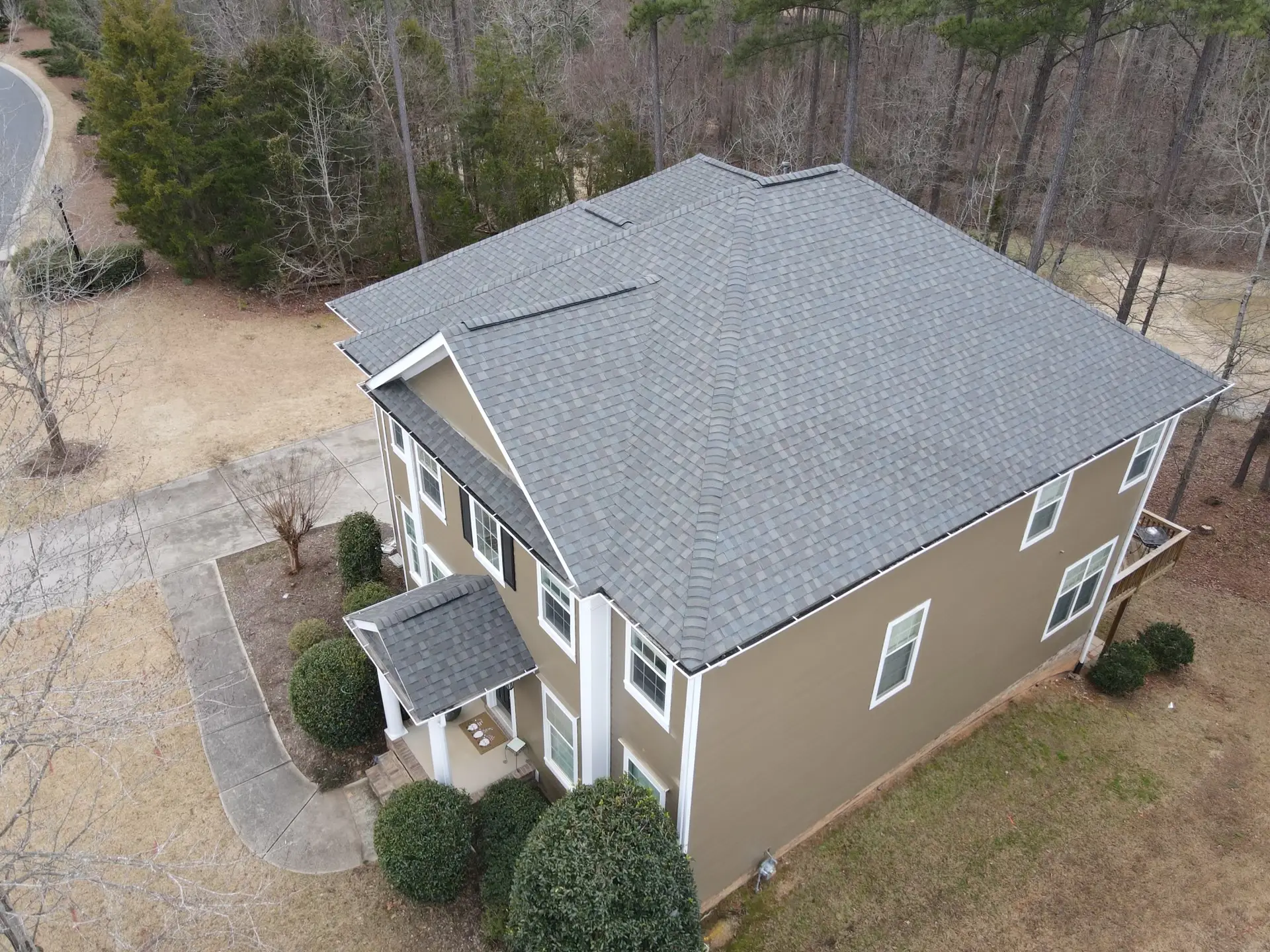 High-angle view of a tan house with white trim and a fresh dark gray asphalt shingle hip roof with multiple ridge vents and a small porch.