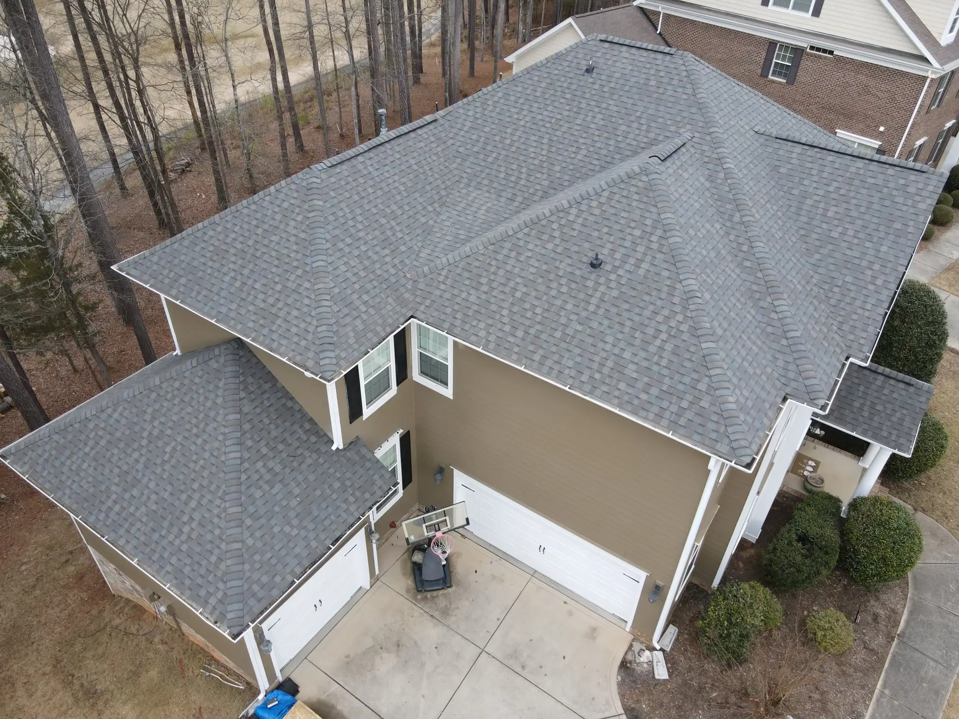 High-angle view of a tan house with white trim and a new dark gray asphalt shingle hip roof with multiple ridge vents and an attached garage.