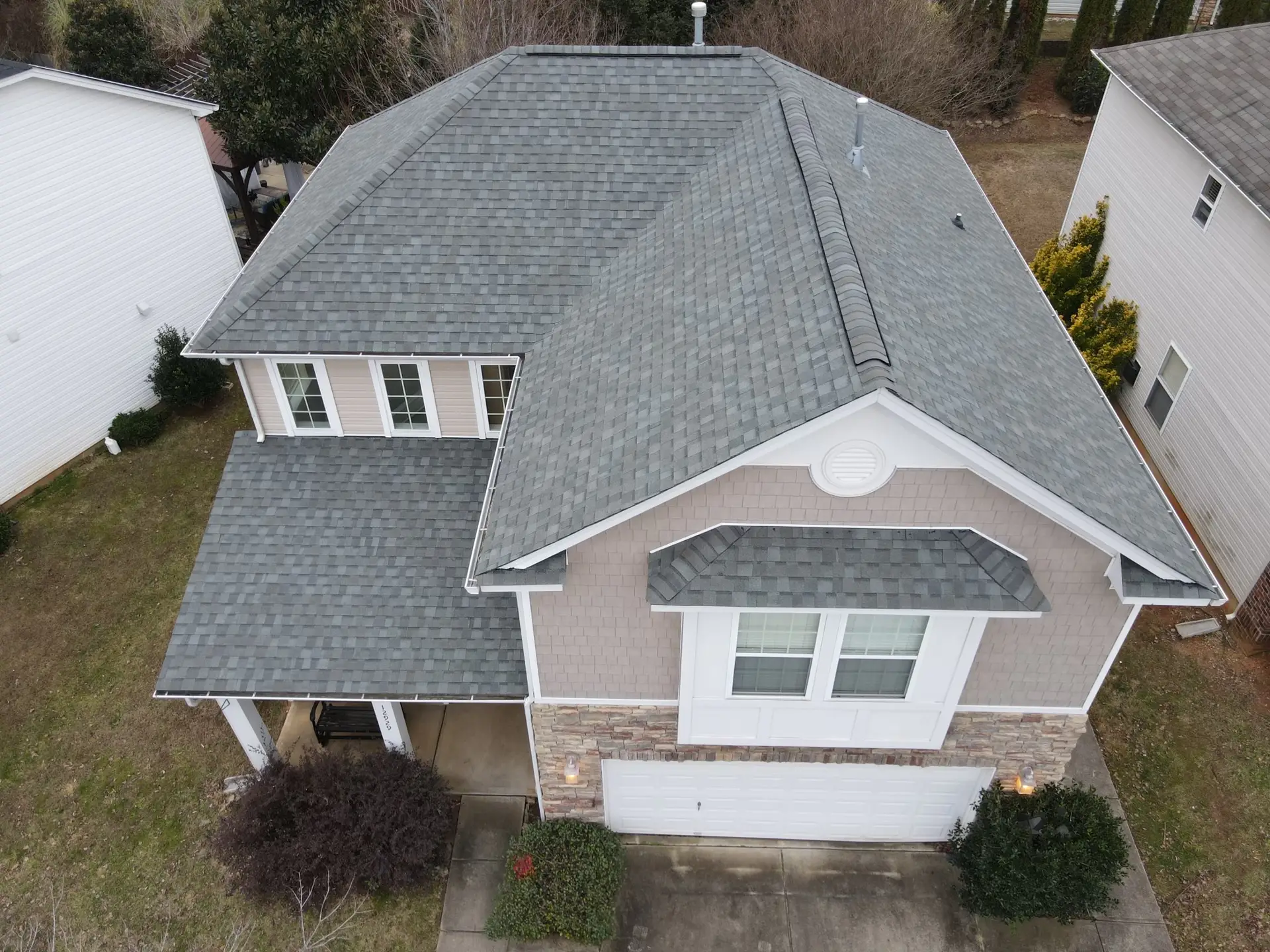 Drone photo showing a beige two-story home with stone siding, white trim, and a newly installed light gray asphalt shingle gable roof.