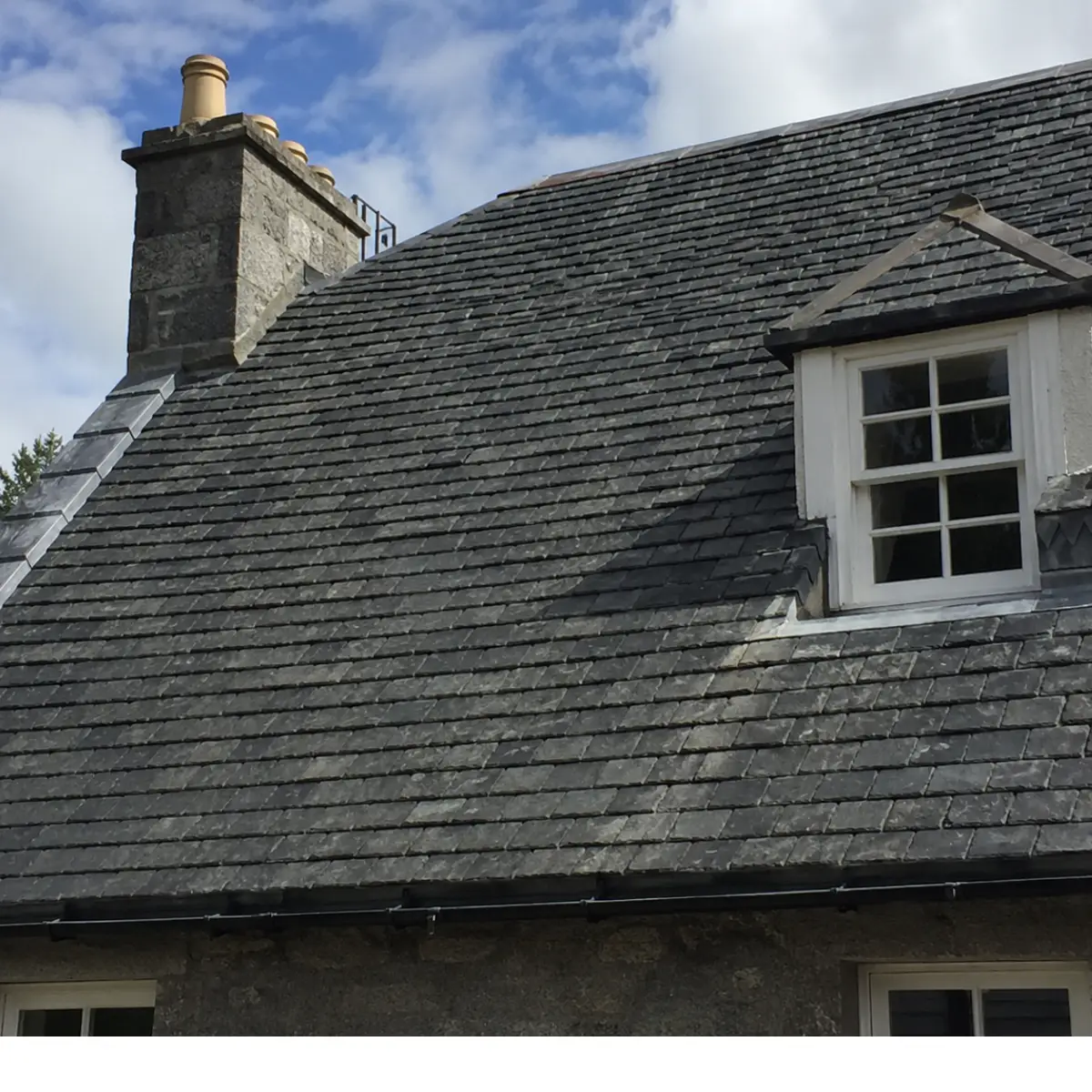 Gray slate roof on a stone house with a stone chimney and dormer window, set against a blue sky