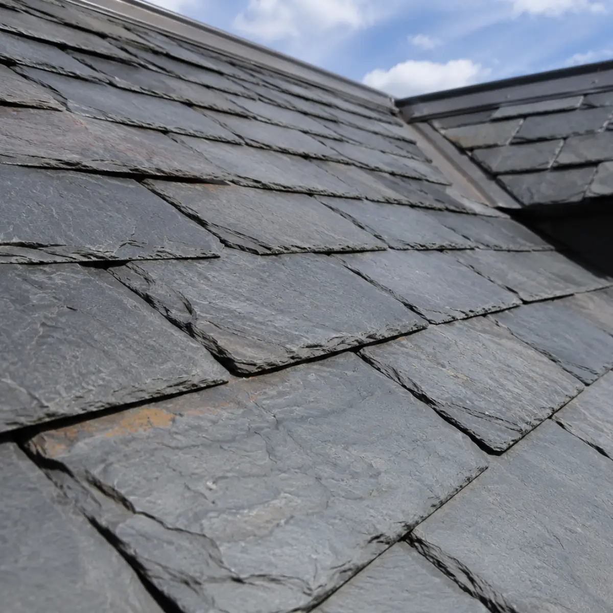 Close-up of natural gray slate roof tiles with visible texture and layered edges, against a blue sky